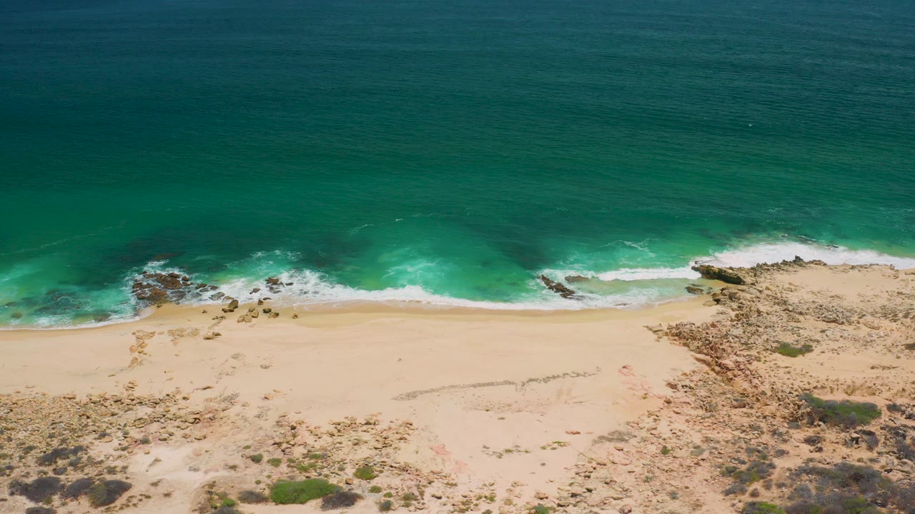 Aerial view of turquoise water, white surf, pale sand, rocks, and dune scrub.