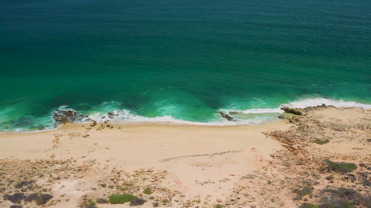 A second San Jose Del Cabo aerial frame shows blue-green water curling onto light sand, with scattered rocks at the left and right edges and no visible people.