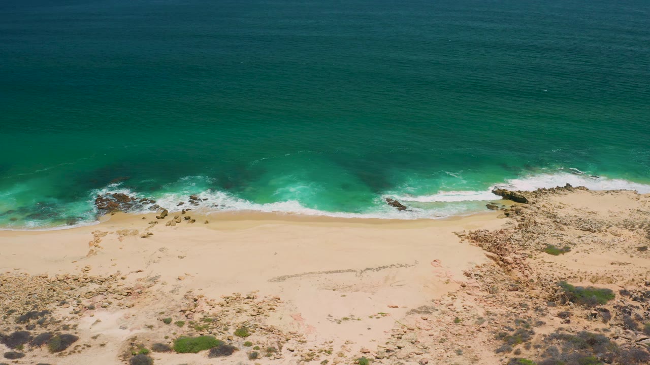 A San Jose Del Cabo shoreline frame shows clear blue water, a white foam band, broad pale sand, rough rocky ground, small green scrub patches, and no visible people.