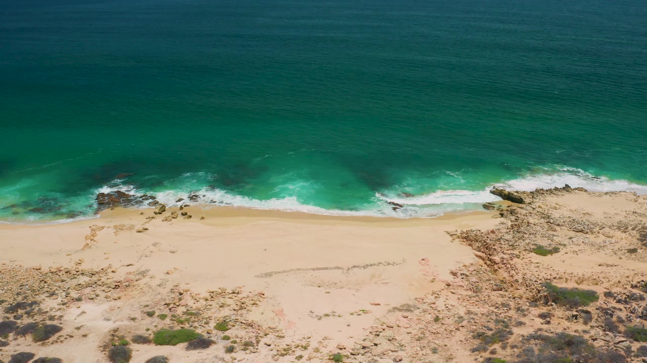 At 0:03, the San Jose Del Cabo beach appears from above with darker teal ocean in the upper frame, surf crossing the center, pale sand, rock clusters, scrub, and no visible people.