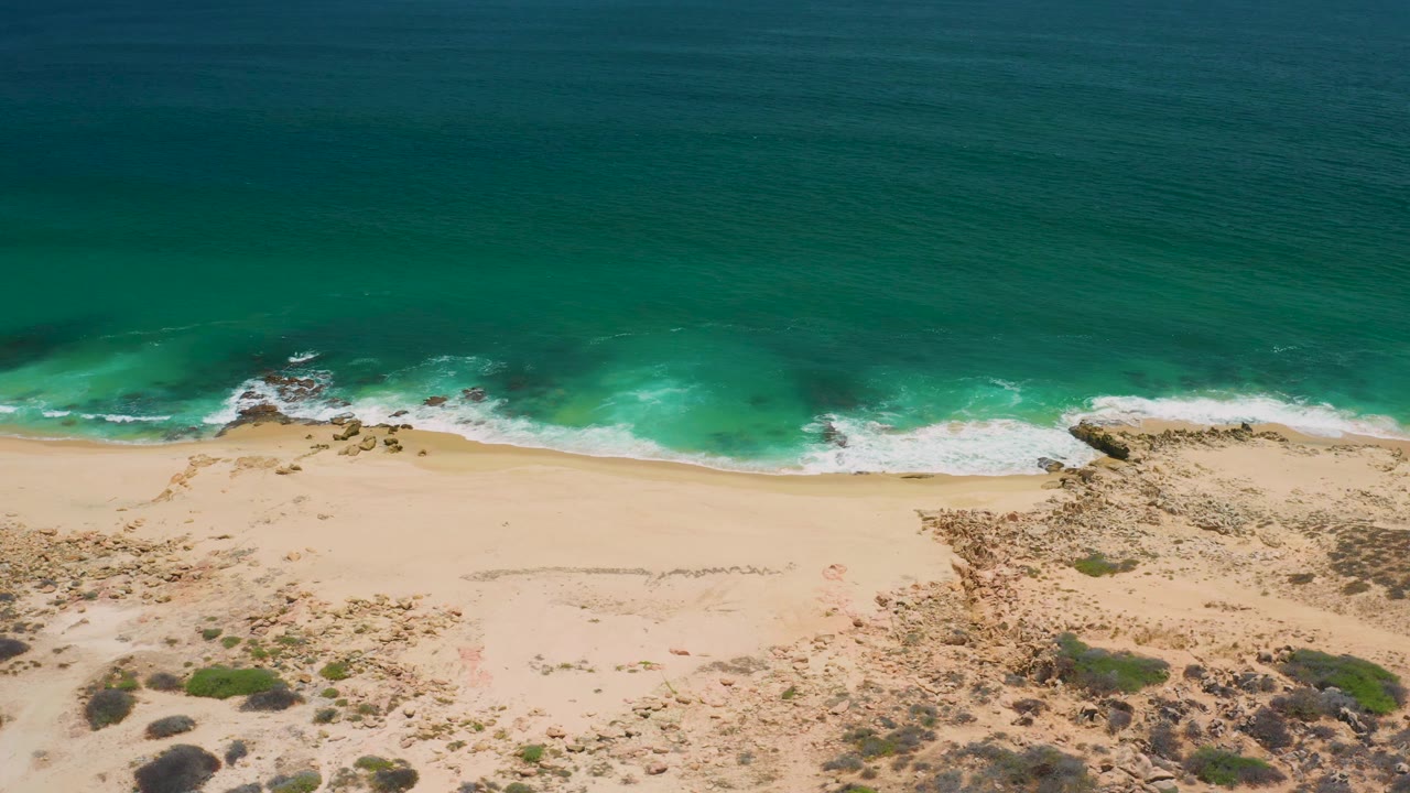Aerial frame of San Jose Del Cabo shoreline where white foam gathers around a rocky patch, with pale sand and scrubland below and no visible people.