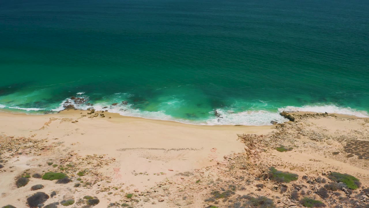 San Jose Del Cabo aerial shoreline frame with a broad band of green water, thin whitecaps, open sand, scattered scrub, and no visible people.