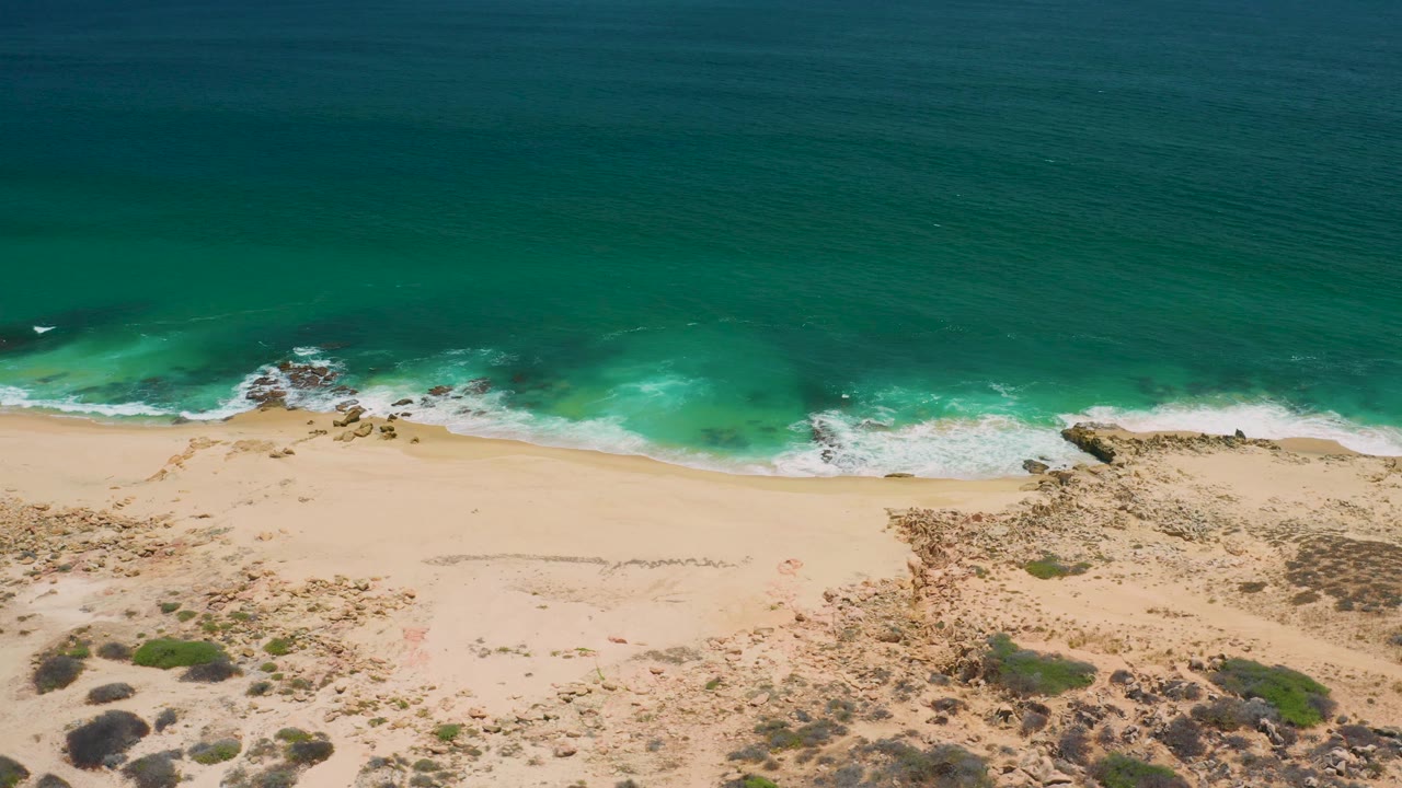 Aerial view over San Jose Del Cabo surf, with darker green ocean at the top, a white foam edge, pale sand, scrub patches, and no visible people.