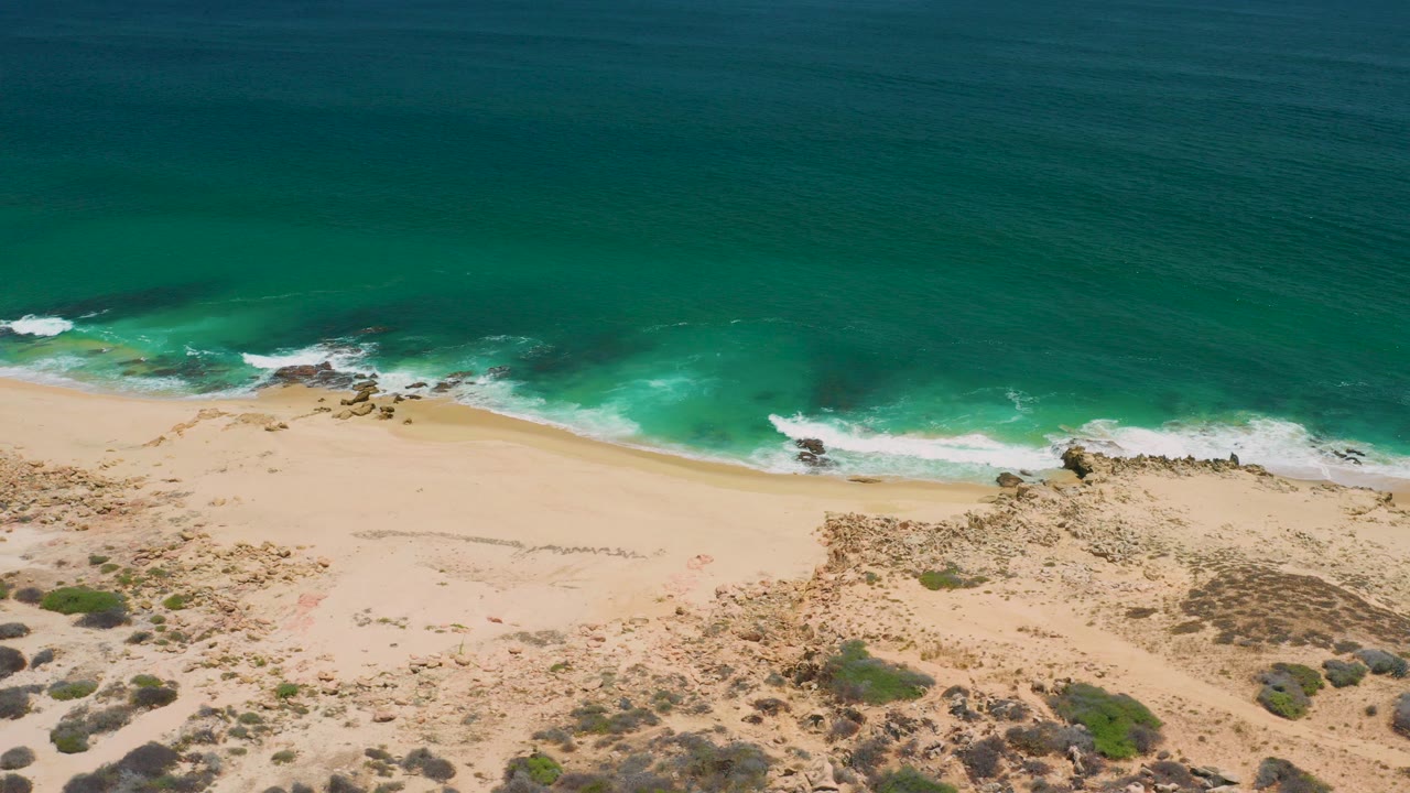 Aerial view in San Jose Del Cabo, Mexico, with deep turquoise water at the top, white surf along pale sand, dry scrub in the foreground, and no clearly recognizable people.