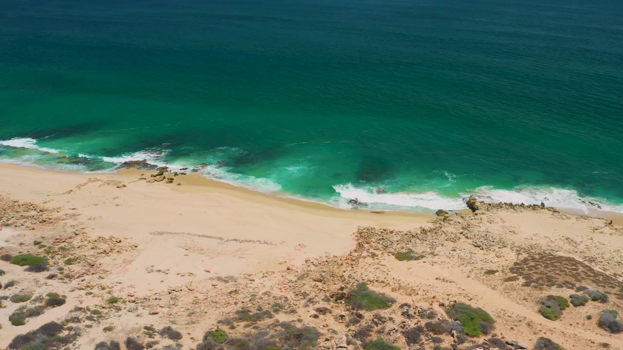 High aerial view in San Jose Del Cabo, Mexico, with broad turquoise water, white foam near shore, a sandy beach band, rocky outcrops, and scrub-covered dunes in the lower frame.
