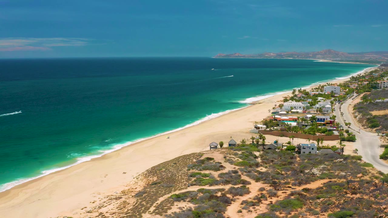 Opening aerial frame over San Jose del Cabo, Mexico with turquoise ocean on the left, pale sand below, low shoreline homes to the right, distant land on the horizon, and no visible people.