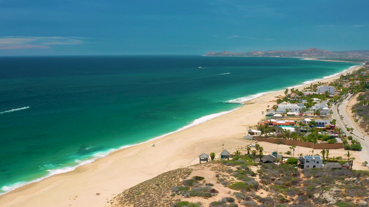 San Jose del Cabo aerial still with turquoise water meeting pale sand, clusters of beachside homes on the right, a curving road beyond them, distant coast on the horizon, and no visible people.