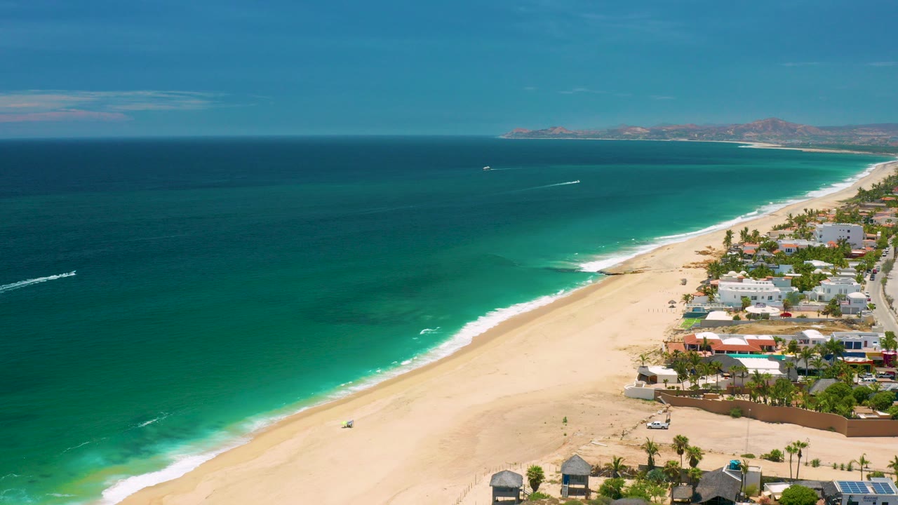Aerial frame over San Jose Del Cabo, Mexico showing green ocean, a diagonal sandy beach, shoreline homes at right, distant mountains, boat wakes, and no visible people.