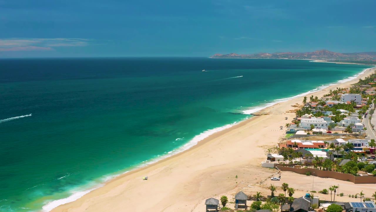 Green surf meets a wide beach with buildings and palms along the right edge.