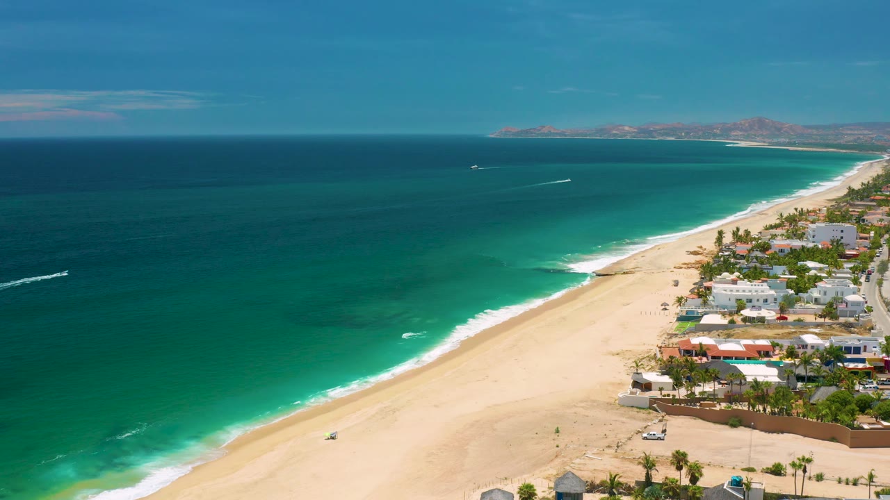 High aerial view in San Jose Del Cabo, Mexico with turquoise shallows, a smooth sandy beach, white homes near palms, mountain ridges in the distance, and no visible people.