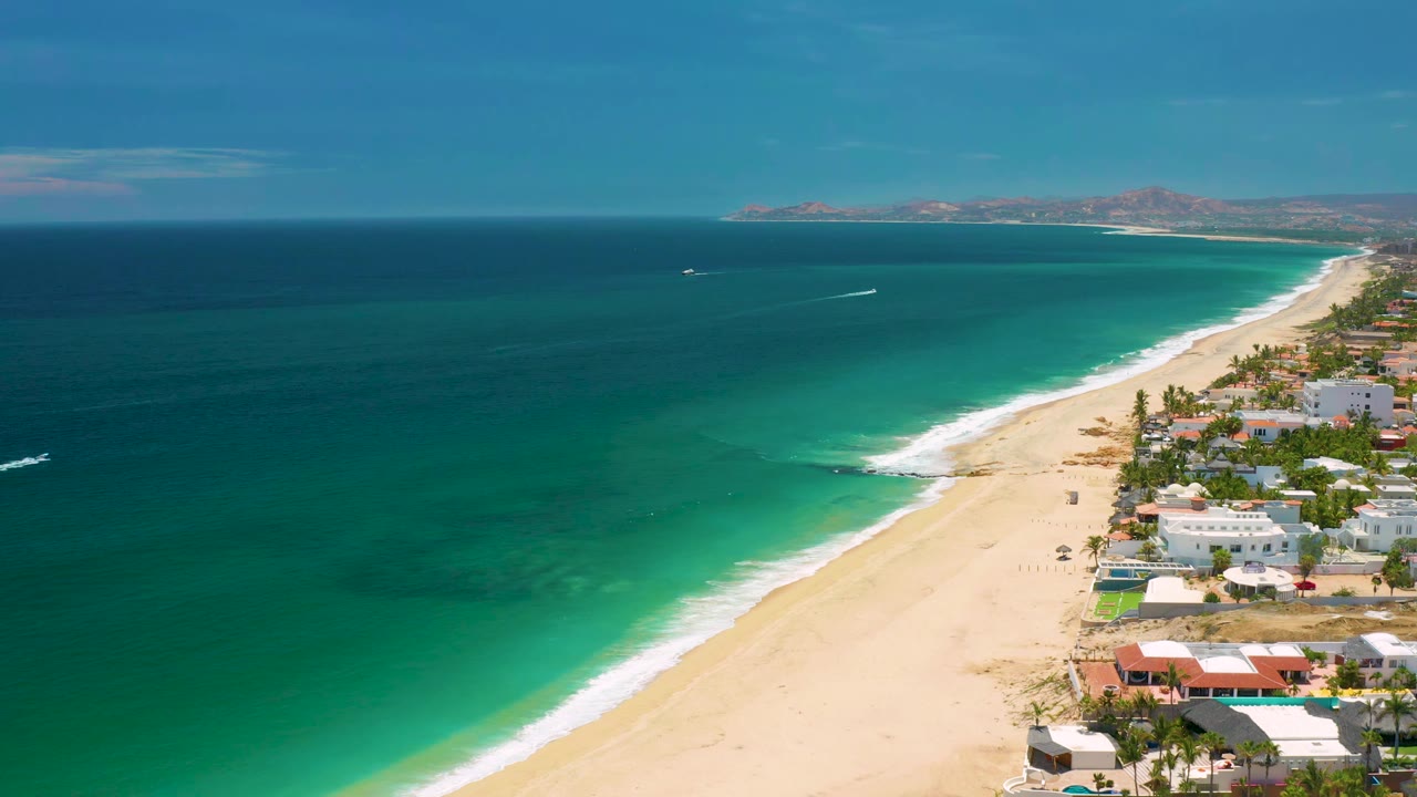 Drone view of blue ocean beside a long sandy beach and coastal buildings.