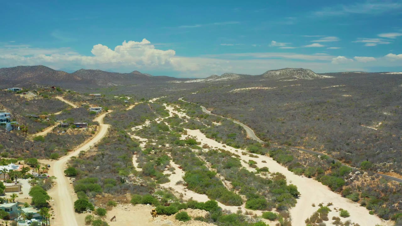 High aerial frame over San Jose Del Cabo terrain with a winding dirt road, scrub-covered slopes, distant ridges, scattered hillside buildings, and no visible people.