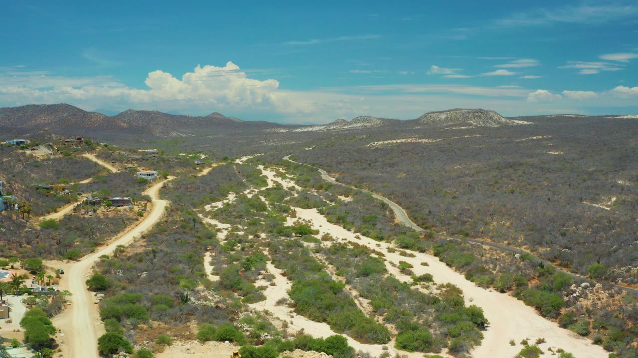 Aerial frame over San Jose Del Cabo dry hills with pale sandy washes, winding dirt trails, scattered scrub, distant mountains, blue sky, and no visible people.