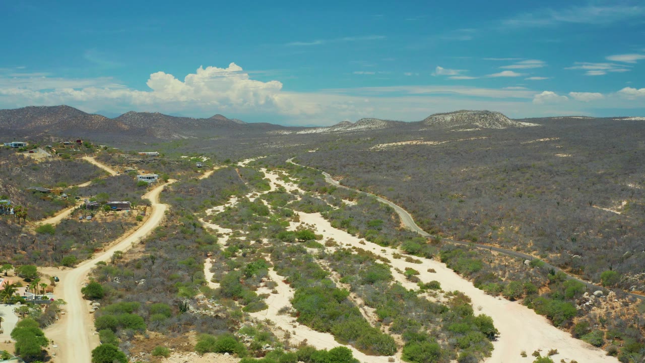 Drone frame over San Jose Del Cabo dry valley terrain with branching pale tracks, dark scrub patches, rounded mountains on the horizon, clear sky, and no visible people.