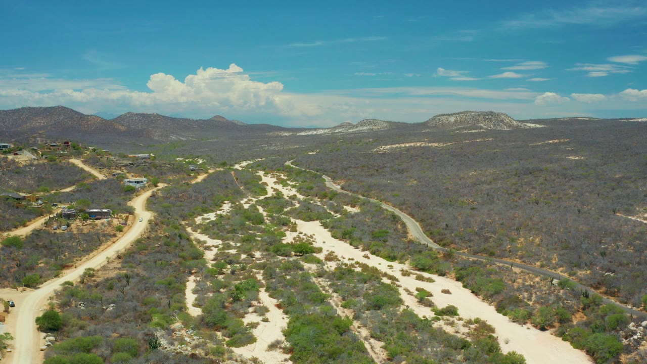 Aerial view over San Jose Del Cabo desert terrain with pale sandy washes, sparse green shrubs, dirt roads, distant hills, and no visible people.