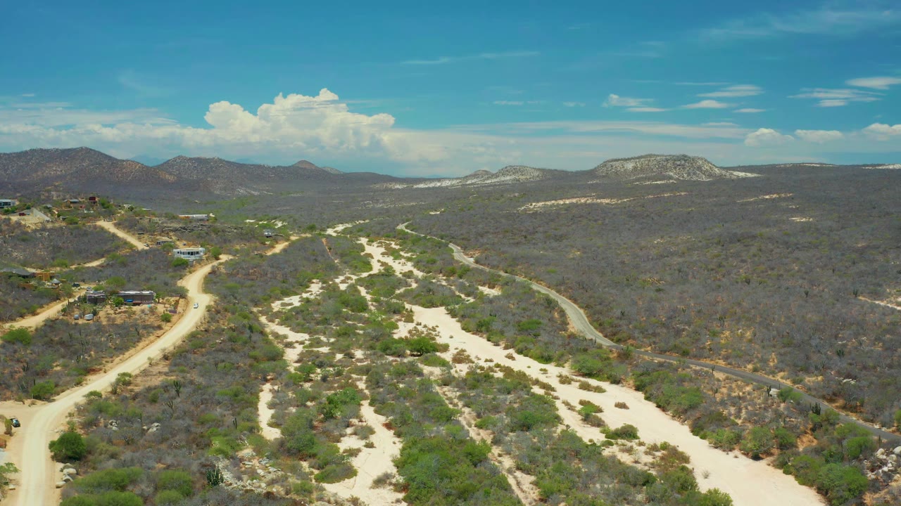 Drone view in San Jose Del Cabo showing a branching pale sand wash through scrubland, a dirt road at left, distant ridges, hillside structures, and no visible people.