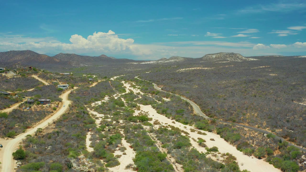 Wide San Jose Del Cabo aerial frame with sandy wash beds in the foreground, scrub-covered hills in the midground, a mountain horizon, small buildings, and no visible people.