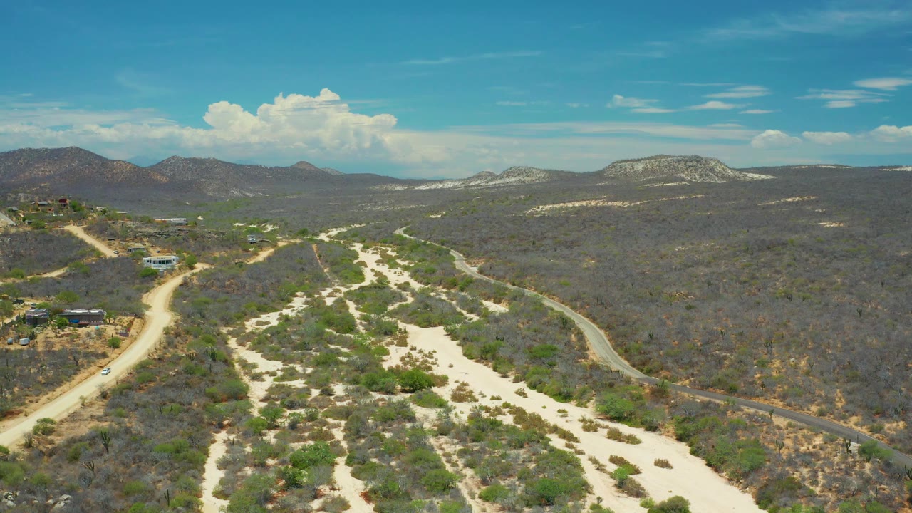 Wide San Jose Del Cabo aerial with a pale dry riverbed in the foreground, a winding road to the right, scattered development to the left, distant mountains, and no visible people.
