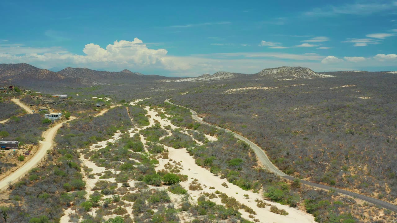 Second frame shows San Jose Del Cabo, Mexico from above, with the sandy wash centered, a dirt road along the left side, small hillside buildings, distant mountains, and no visible people.