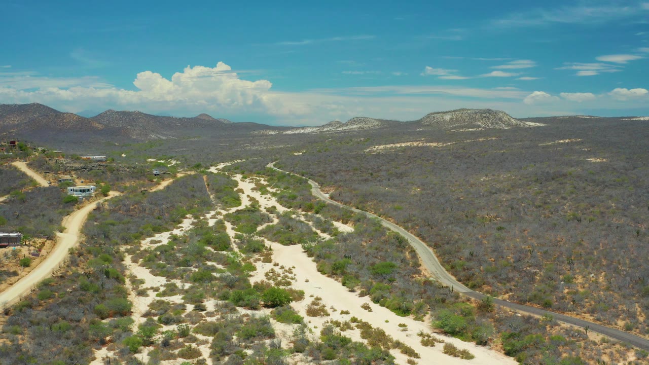 Third frame holds a high view of San Jose Del Cabo, Mexico, with pale sand channels through scrub, a curving road at right, scattered structures at left, distant hills, and no visible people.