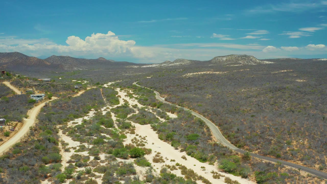 High landscape view with sandy foreground, brush, winding road, hills, and clouds.