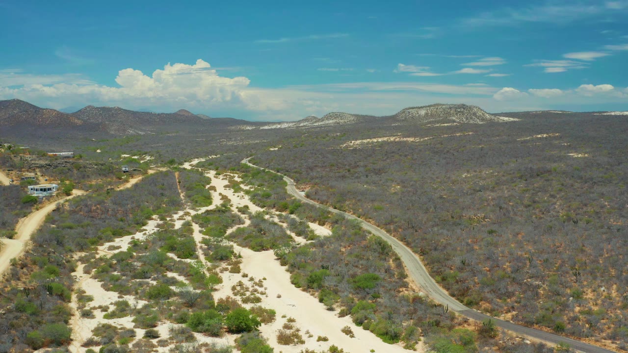 High aerial frame in San Jose Del Cabo showing a pale wash centered in scrubland, a road curving along the right side, low mountains in the distance, and no visible people.