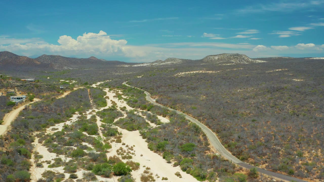 Aerial view over San Jose Del Cabo with branching sandy channels, sparse green shrubs, a narrow road at frame right, distant mountain ridges, and no visible people.