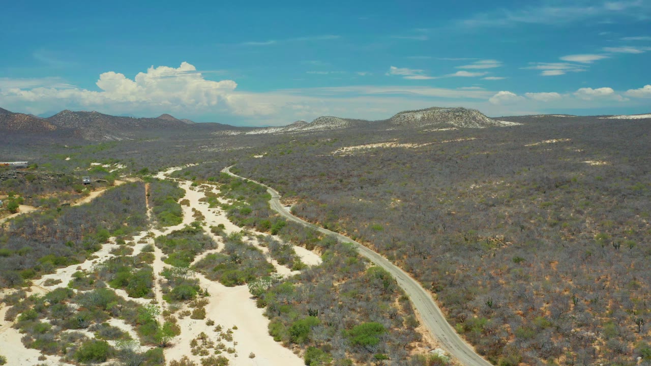 Aerial view in San Jose del Cabo, Mexico, with branching sandy channels below, the road tracking along the right edge, scrub-covered flats, mesa ridges, cloud formations, and no visible people.