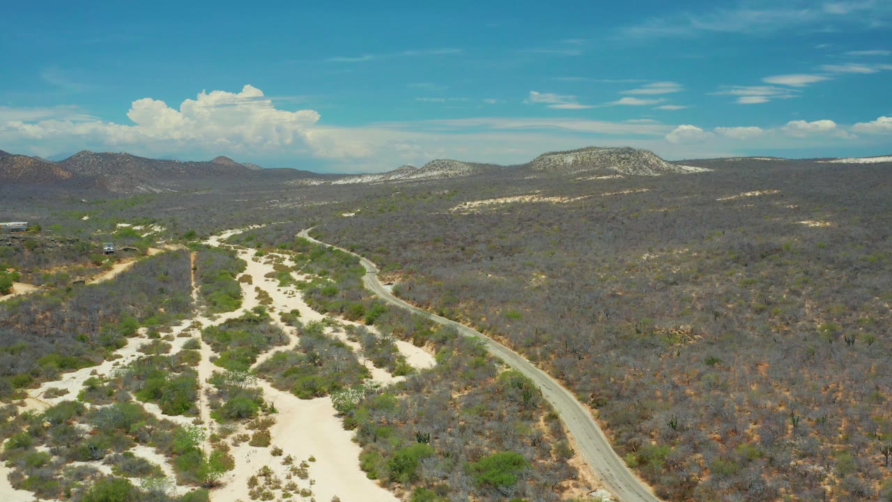 Wide drone frame over San Jose del Cabo, Mexico, showing the road curving through dry brush at right, pale washes at left, dark scrubland across the middle distance, low mountains, and no visible people.