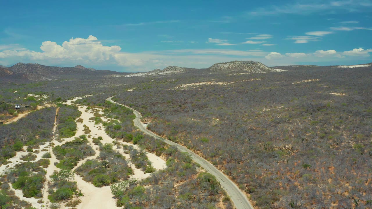 A second aerial frame in San Jose Del Cabo, Mexico shows the curved road closer to the lower edge, with pale sand tracks, scrub vegetation, rocky hills, and no visible people.