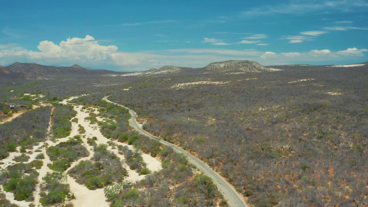 Aerial landscape in San Jose Del Cabo, Mexico with sandy channels running through scrubland, a road along the right side, layered rock formations, clouds, and no visible people.