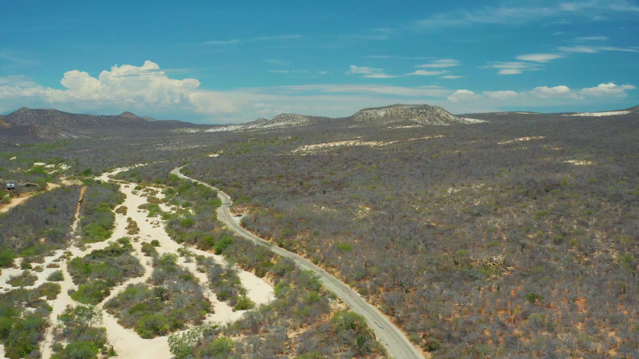San Jose Del Cabo, Mexico aerial frame with sandy pathways spreading across the lower left, a paved road curving through scrub, rocky ridges on the horizon, and no visible people.