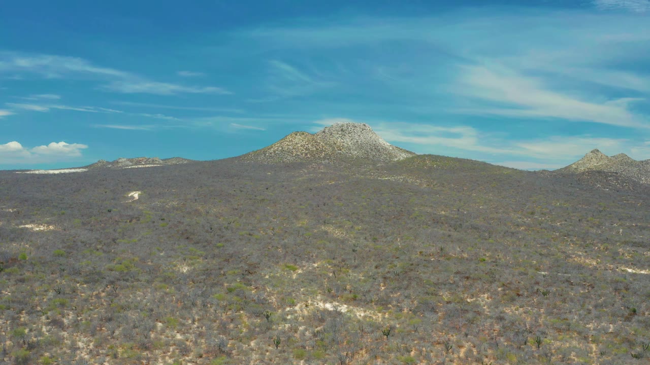 A broad San Jose Del Cabo landscape shows muted green and brown brushland, pale rock patches, and rounded peaks along the horizon with no visible people.