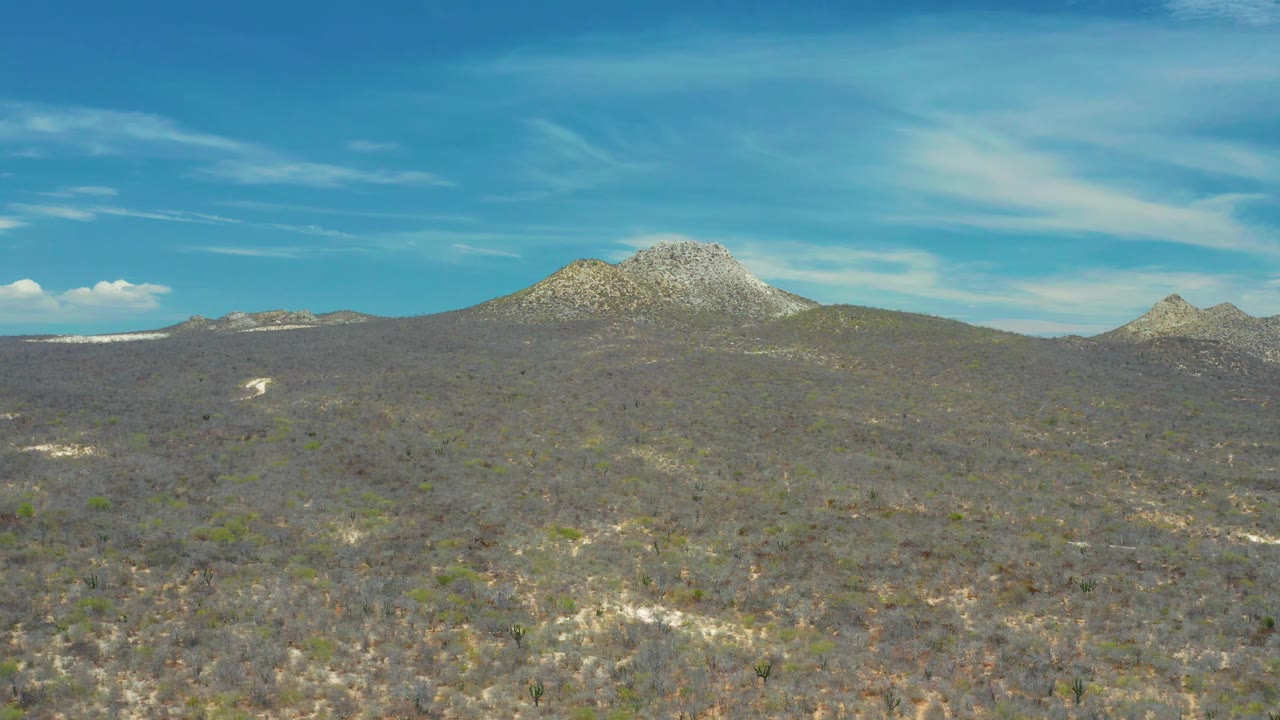 The San Jose Del Cabo frame shows rocky hill country spread across the foreground, a larger peak near the horizon, and thin cloud bands overhead with no visible people.