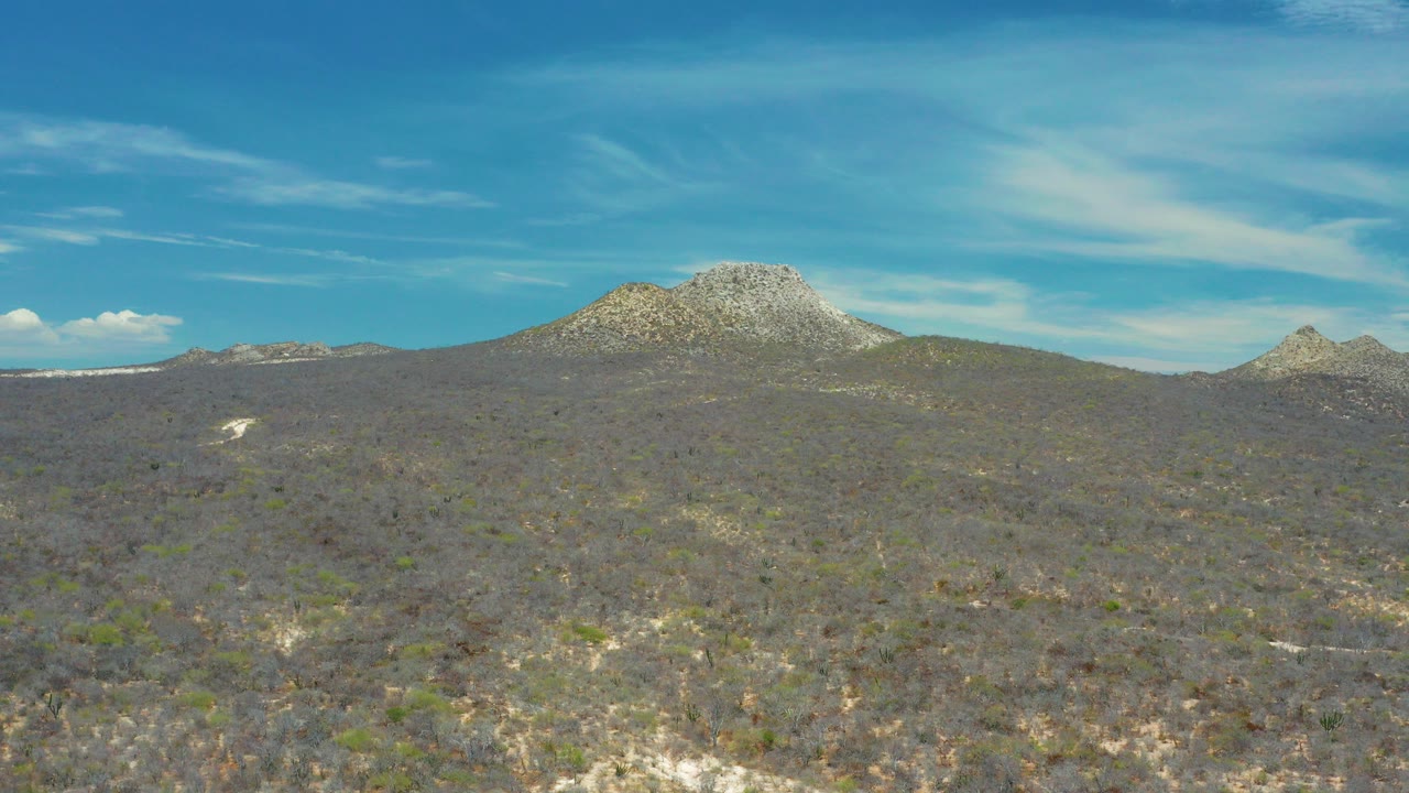 Wide view of rocky scrubland and rounded mountains near San Jose del Cabo, Mexico, with thin clouds in a blue sky and no visible people.