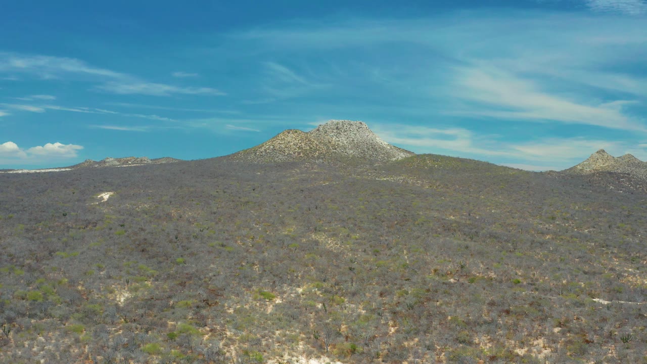 Dry hillside terrain leads to a rounded mountain peak near San Jose del Cabo, Mexico, with wispy cloud streaks across the bright sky and no visible people.
