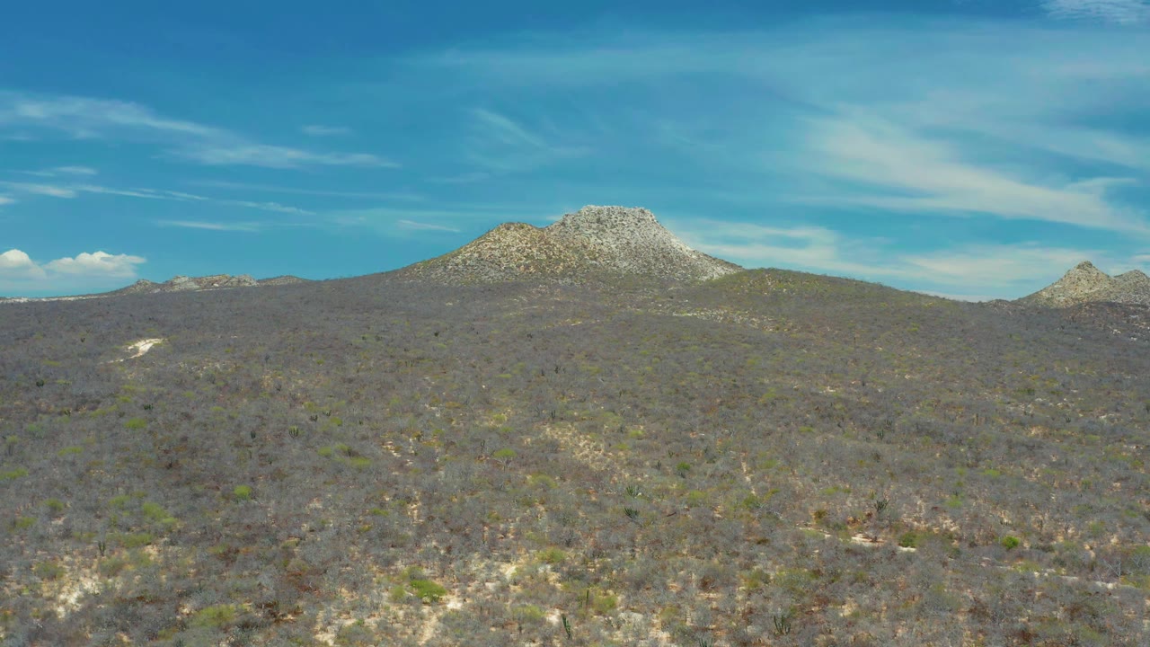 Rocky ridge and dry brushland fill the San Jose Del Cabo landscape, with wispy white clouds spread across the blue sky and no visible people.