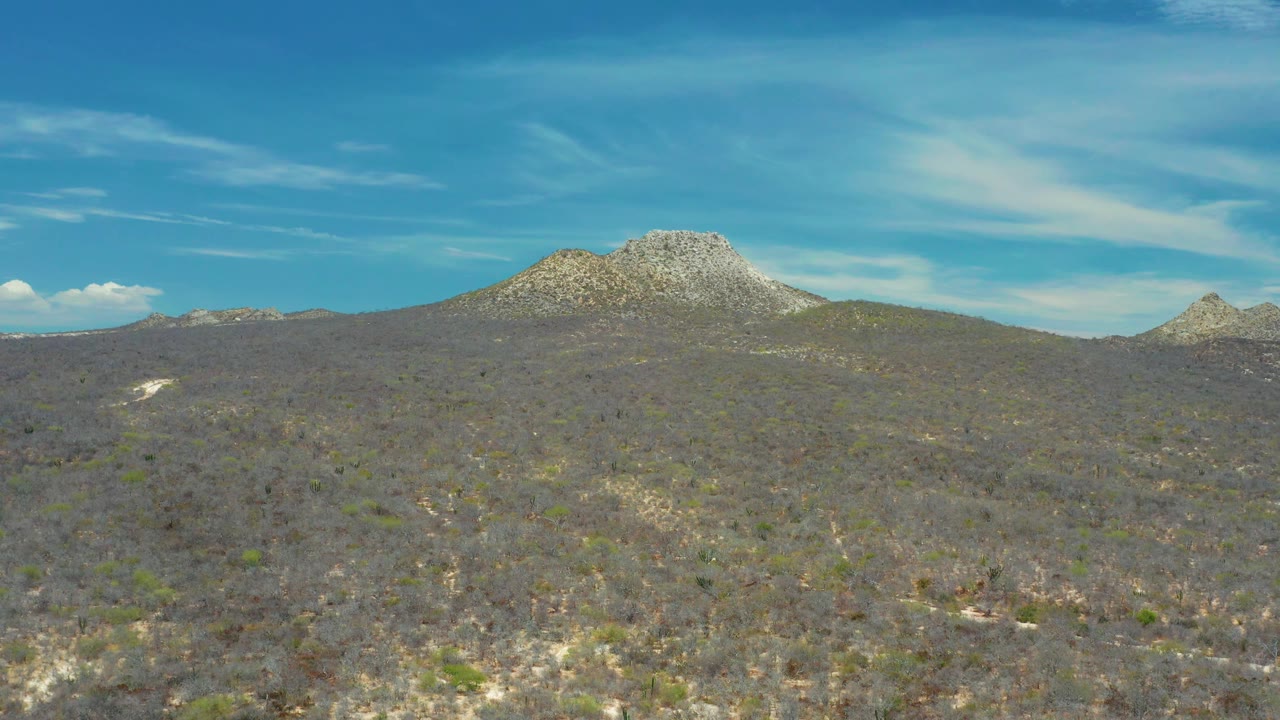 Scrub-covered terrain stretches toward pale rocky peaks near San Jose Del Cabo, Mexico, with streaked clouds high in the blue sky and no visible people.
