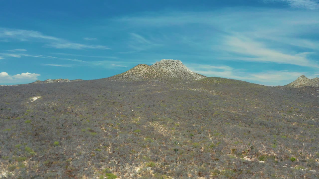 Dry hillside brush leads to a rocky mountain form in the San Jose Del Cabo area, with bright blue sky, white clouds, and no visible people.
