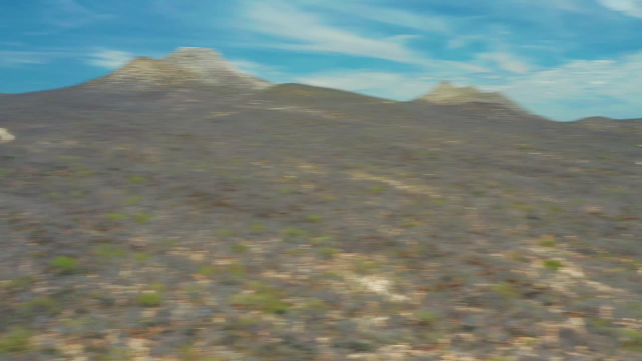 Motion-blurred aerial frame over San Jose Del Cabo scrubland with low desert hills, distant mountains, blue sky, and no visible people.