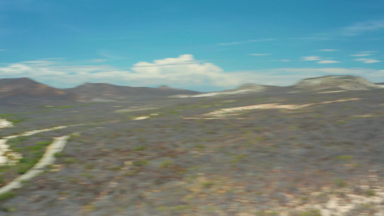 Opening frame of San Jose Del Cabo aerial terrain with dry scrubland, pale washes, distant hills, motion blur, and no visible people.