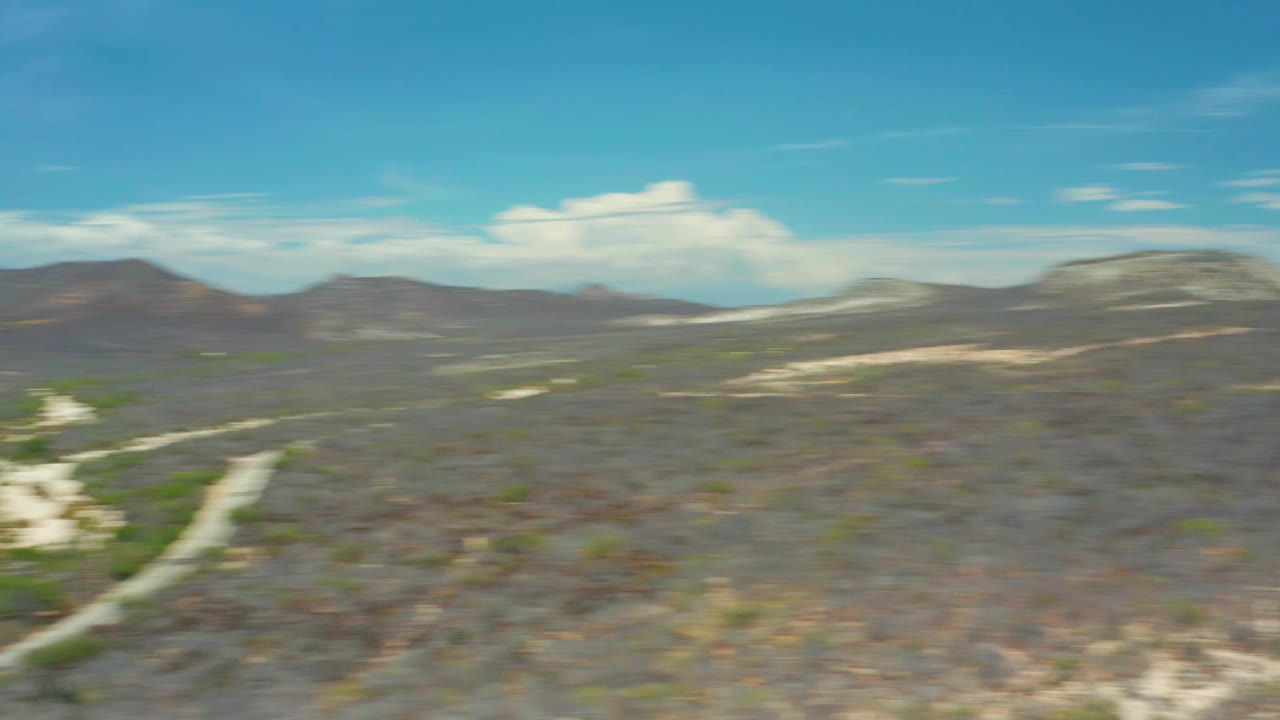 Second frame keeps the San Jose Del Cabo valley broad in view, with dry brush, a pale wash, low mountain ridges, blue sky, and no visible people.