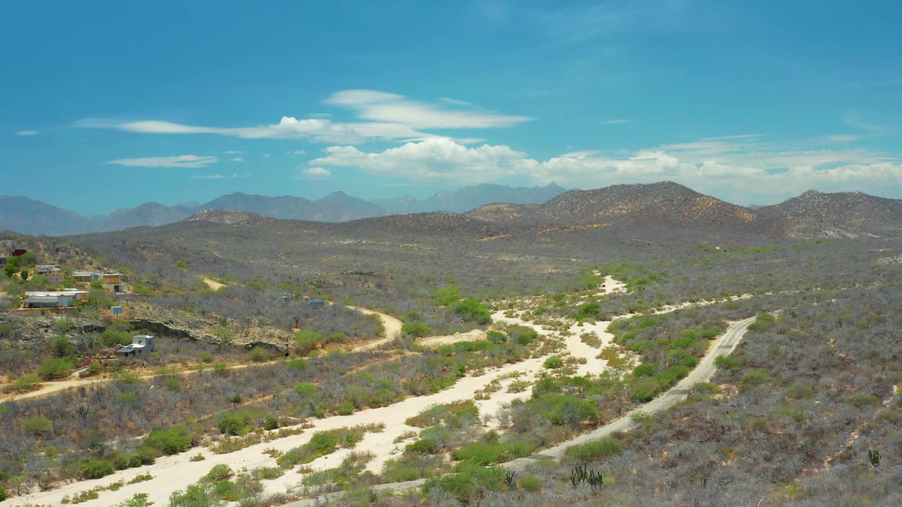 San Jose Del Cabo aerial view with scattered small buildings, winding dirt roads, dry scrub hills, distant mountains, light clouds, and no visible people.
