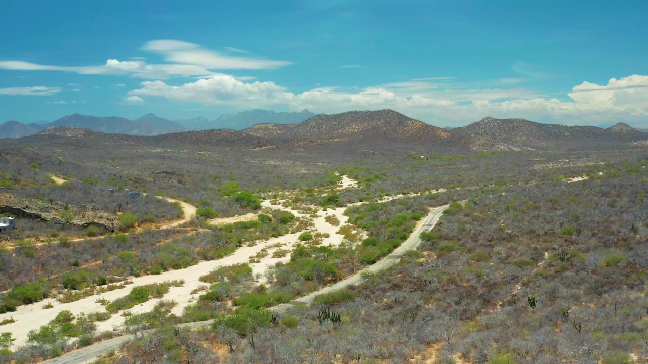 Aerial frame from San Jose Del Cabo, Mexico showing pale dirt roads through dry scrubland, low hills, distant blue mountains, scattered clouds, and no visible people.