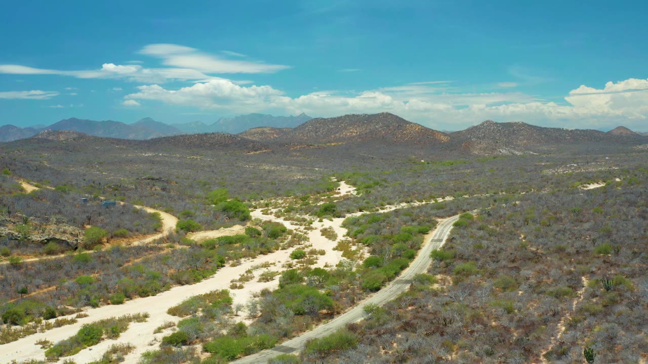 Wide aerial frame in San Jose del Cabo, Mexico, showing the pale wash and roadside scrub shifting across the lower frame with low mountain ridges beyond, with no visible people.