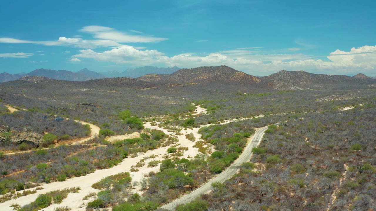 Aerial view in San Jose del Cabo, Mexico, with a narrow road beside a pale sandy channel, scrub-covered hills, distant mountains, and scattered clouds, with no visible people.