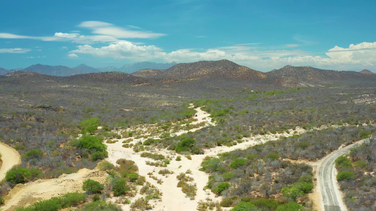 High aerial frame of San Jose del Cabo arid terrain with pale washes crossing gray-green brush, low rounded hills, distant blue mountains, and no visible people.