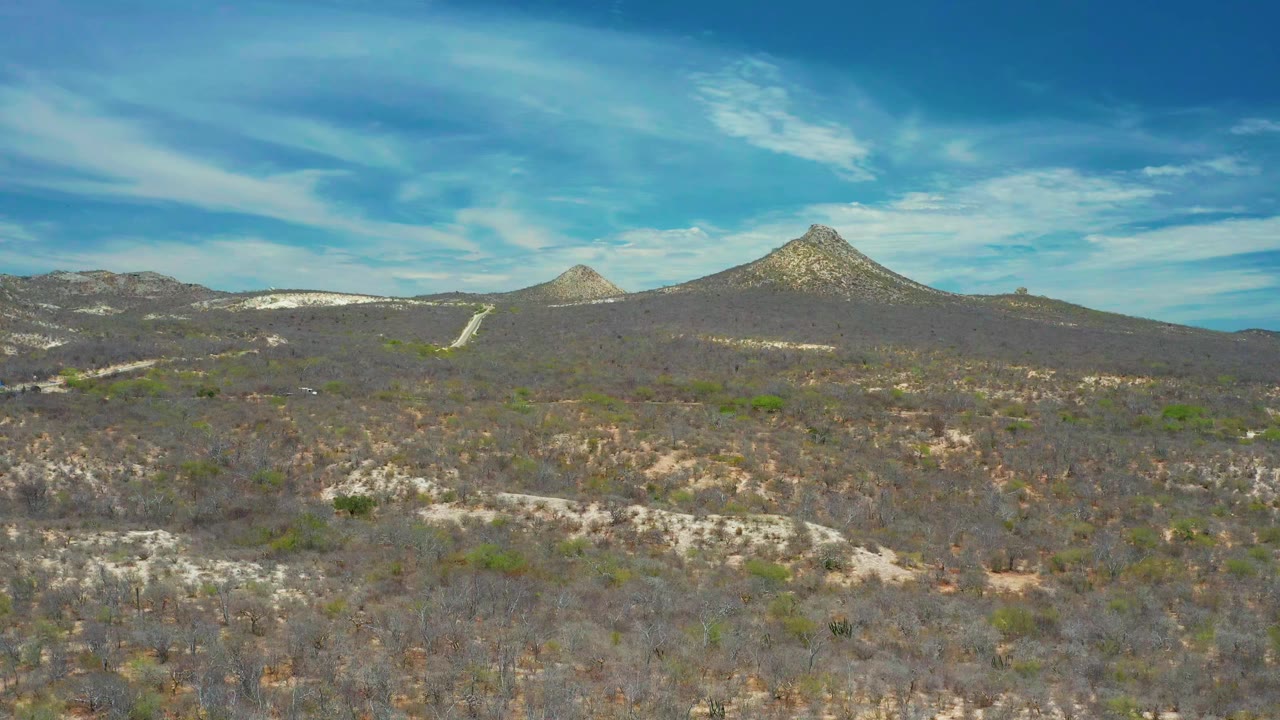 Aerial frame of San Jose del Cabo desert scrub with a pale road or track crossing the midground below pointed hills, with no visible people.
