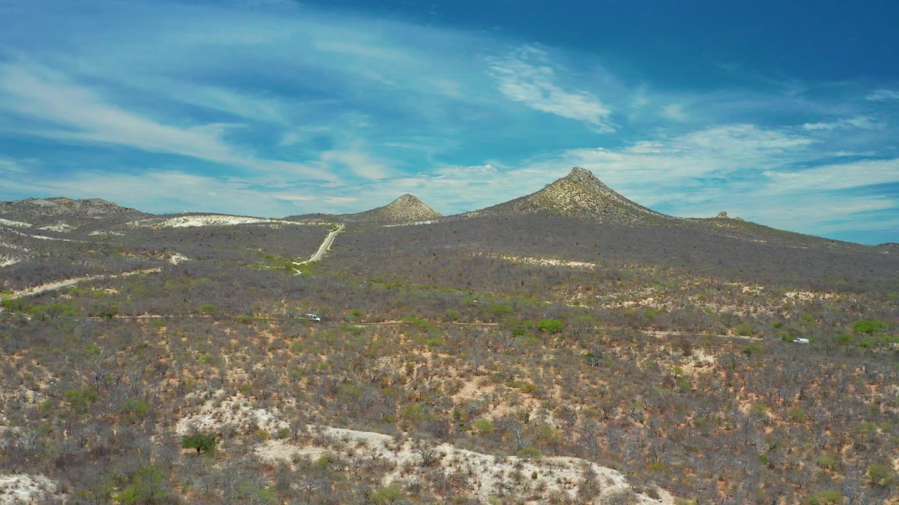 Wide elevated view of dry scrub hills and a pointed mountain near San Jose Del Cabo, Mexico, with a pale road below and no visible people.