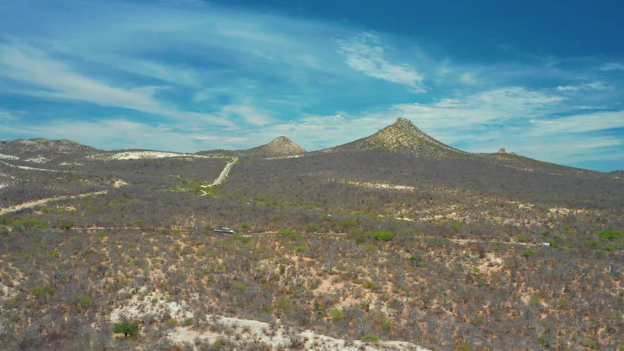 Wide 4K aerial frame of San Jose Del Cabo scrubland with a lower road, small vehicles, pale hillside cuts, a rocky mountain ridge, and no visible people.
